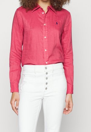 Žena v červené košili s dlouhým rukávem zapnuté na knoflíky, zastrčené do bílých kalhot s vysokým pasem a několika předními knoflíky.