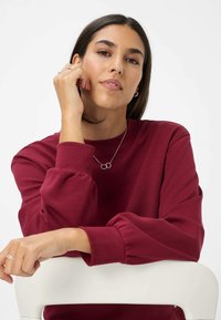 Bordeaux sweatshirt met lange mouwen, met een relaxte pasvorm en een pofmouwontwerp. De persoon draagt zilveren sieraden en poseert met een hand onder hun kin.