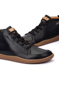 Un par de zapatos casuales de cuero negro con suelas de goma marrón y cordones negros, angulados para mostrar detalles del lateral y la parte frontal.