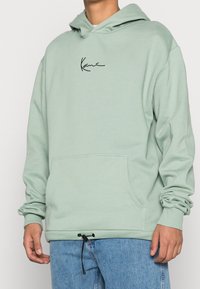 Felpa verde menta con tasca frontale, cappuccio con lacci e dettagli del logo neri. Realizzata in tessuto morbido con polsini e orlo a coste.