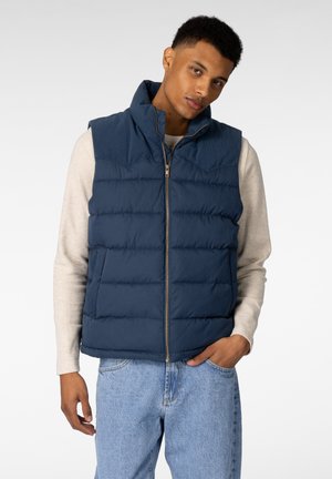 Mørkeblå dunvest med høy krage, frontglidelås og kanalsøm. Brukt over en lys beige langermet skjorte, kombinert med jeans.