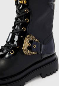 Botas tobilleras negras con cordones y acabado brillante de charol, que presentan una hebilla decorativa dorada y una suela de goma gruesa con textura.