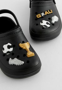Zuecos negros con varios charms de Jibbitz de temática deportiva: balón de fútbol, silbato, trofeo y zapato de fútbol, con agujeros de ventilación en toda su superficie.
