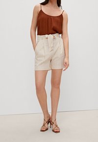 Top sans manches marron avec un col froncé, associé à un short taille haute beige à rayures avec une ceinture nouée. Talons hauts à lanières marron.