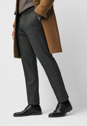 Hiilenharmaat ruudulliset housut yhdistettyinä mustiin nahkakenkiin ja ruskeaan villakangastakkiin. Leikkaus on kapea ja materiaali hienostuneesti teksturoitu.