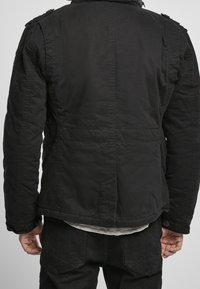 Muž s černou bundou a černými kalhotami, ukázaný zezadu s uvolněnýma rukama podél těla na hladkém pozadí.