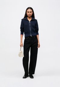 Námornícky zipsom zapínaný pletený cardigan s vysokým golierom, červenými a bielymi pruhmi; skombinovaný s čiernymi širokými nohavicami a krémovou kabelkou.