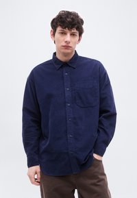 Jeune homme aux cheveux bouclés et foncés portant une chemise en velours côtelé bleu marine avec une poche poitrine et un pantalon marron, debout sur un fond blanc uni.