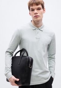 Ανοιχτό πράσινο πόλο με μακριά μανίκια και μικρό λογότυπο· συνδυάζεται με μια μαύρη δερμάτινη τσάντα με χερούλια στο πάνω μέρος και κλείσιμο με φερμουάρ.