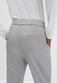 Lichtgrijze broek van een gestructureerde stof, met een verborgen achterzak en een platte tailleband, gecombineerd met een witte blouse met lange mouwen.
