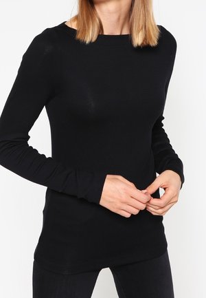 Kobieta ubrane w dopasowaną czarną koszulę z długim rękawem, stojąca z rękami luźno splecionymi na wysokości talii na białym tle.