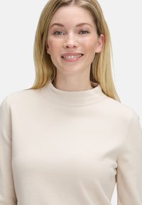 Glimlachende vrouw met lang blond haar, gekleed in een lichtbeige hooggesloten trui, tegen een effen witte achtergrond.