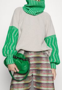 Hellgrauer Pullover mit grün gestreiften Ärmeln und Kragen, kombiniert mit mehrfarbigen strukturierten Hosen und einer grünen Handtasche.