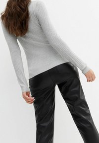 Personne aux longs cheveux bruns portant un pull gris clair moulant à côtes et un pantalon en cuir noir, posant devant un fond blanc uni.