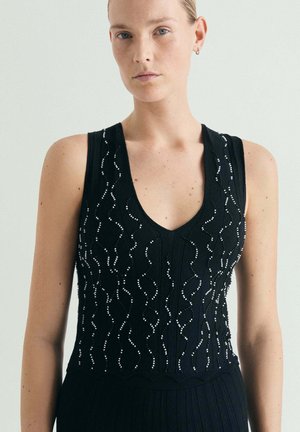 Mujer con una blusa negra sin mangas con patrones ondulados de cuentas y escote en V, de pie frente a un fondo claro y liso.