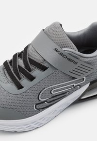 Chaussure de sport grise avec une tige en mesh texturé, lacets noirs et une sangle renforcée. Présente une semelle blanche avec un motif de semelle géométrique.