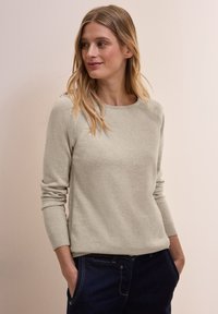 Hellgrauer Pullover mit rundem Halsauschnitt und gerippten Bündchen. Er zeichnet sich durch eine glatte Textur und subtile Raglannaht aus, kombiniert mit dunkelblauen Jeans.