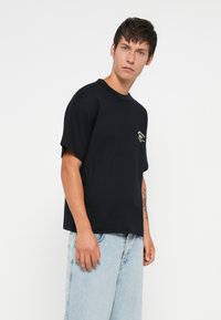 T-shirt nera a maniche corte in cotone, con un piccolo logo sul petto. Il modello indossa jeans in denim blu chiaro con un fit rilassato.