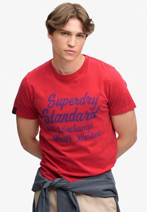 Ung mand iført en rød t-shirt med blå tekst "Superdry Standard" og en blå jakke bundet omkring taljen, stående mod en ensfarvet baggrund.