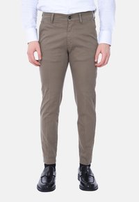 Pantaloni di cotone khaki con vestibilità slim, dotati di chiusura con bottone, passanti per cintura e tasche laterali. Indossati con una camicia bianca a maniche lunghe e scarpe nere.