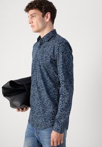 Long-sleeve shirt featuring a navy background with white abstract patterns. Smooth texture, button-up design, paired with a black bag.