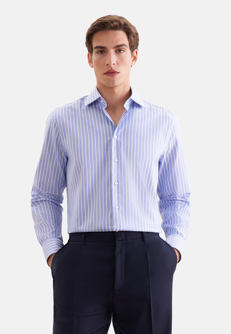 Jeune homme portant une chemise rayée verticale bleu et blanc rentrée dans un pantalon bleu marine foncé, mains dans les poches, regardant vers l'avant.