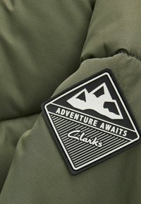 Tessuto imbottito verde oliva con finitura texturizzata, dotato di un badge in gomma nera con grafica di montagna e il testo "L'Avventura Ti Aspetta".