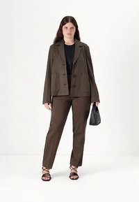 Ruskea, teksturoidusta kankaasta valmistettu bleiseri ja siihen sopivat housut, joissa on hienovarainen kuviointi. Malli pitää kädessään pientä mustaa käsilaukkua ja pukeutunut mustiin remmikkäsandaaleihin.