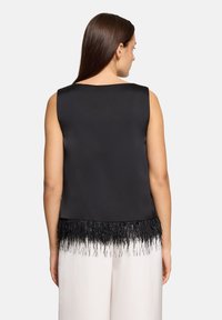 Czarny bezr�ękawnik o gładkim satynowym wykończeniu i frędzlowanym dole z teksturowanymi akcentami, zestawiony z jasnymi spodniami.