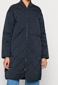 Cappotto trapuntato blu navy con zip frontale, collo a coste e tasca laterale. Caratterizzato da maniche lunghe e orlo arrotondato. Tessuto liscio in tutto il capo.