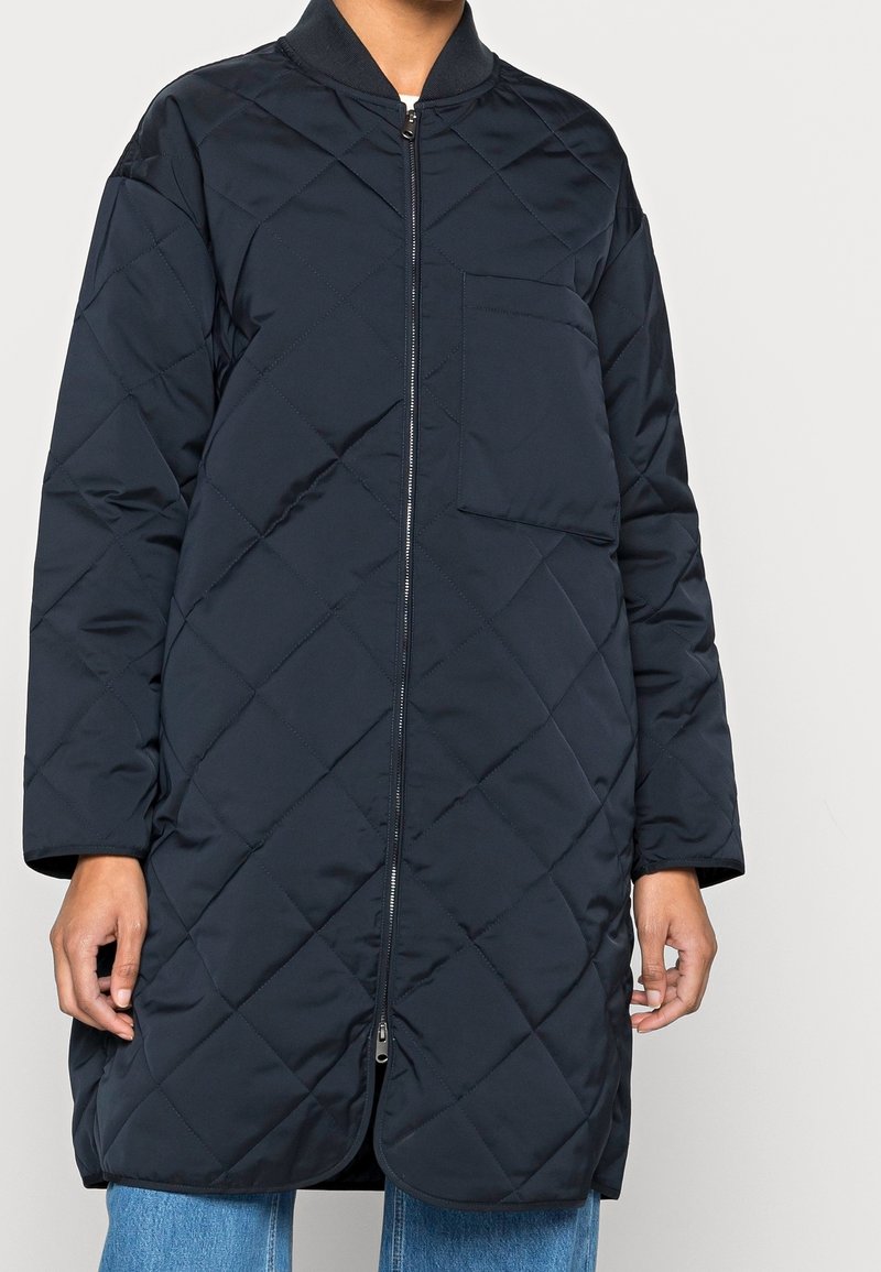 Cappotto trapuntato blu navy con zip frontale, collo a coste e tasca laterale. Caratterizzato da maniche lunghe e orlo arrotondato. Tessuto liscio in tutto il capo.