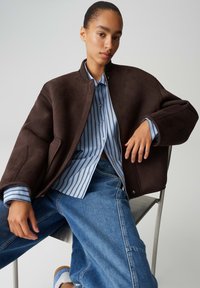 Giacca bomber in camoscio marrone con collo a coste, indossata sopra una camicia a righe azzurre e bianche, abbinata a jeans in denim a vestibilità rilassata.