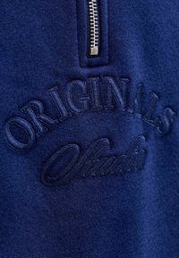 Námořnická modrá mikina se zipem na krku. Obsahuje texturovaný vyšívaný nápis "ORIGINALS" a "Studio" v mírně tmavší modré.