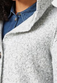 Šedá texturovaná vlněná bunda s širokým límcem, vyrobená z jemných, kudrnatých přízí. Nosená přes modrou džínsovou košili s zapínáním na knoflíky.