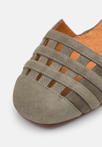 Gros plan d’une chaussure plate en suède taupe avec un bout fermé et un design de bride découpée sur le dessus.