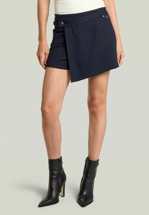 Donna che indossa uno skort nero asimmetrico e stivaletti neri con tacco su uno sfondo chiaro uniforme.