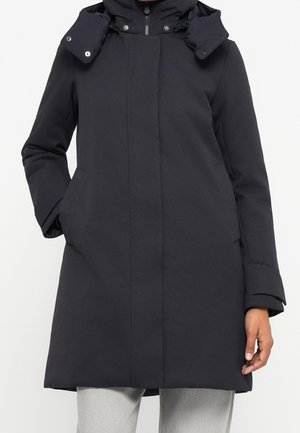 Kobieta w długim czarnym zimowym płaszczu z kapturem, zapinanym z przodu na zamek i wyposażonym w kieszenie na ręce, w połączeniu z jasnoszarymi spodniami.