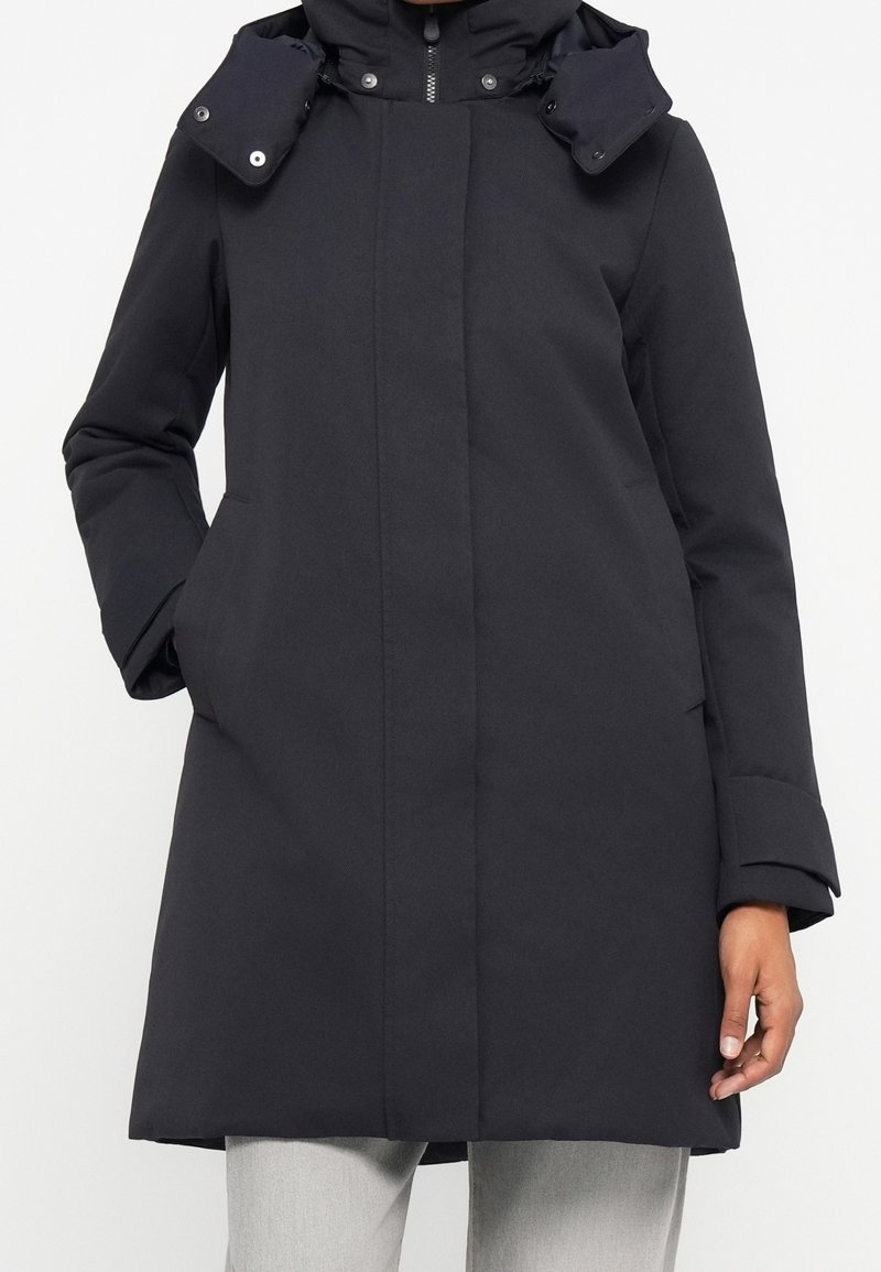 Mujer que lleva un abrigo de invierno largo negro con capucha, cremallera frontal y bolsillos para las manos, combinado con pantalones gris claro.