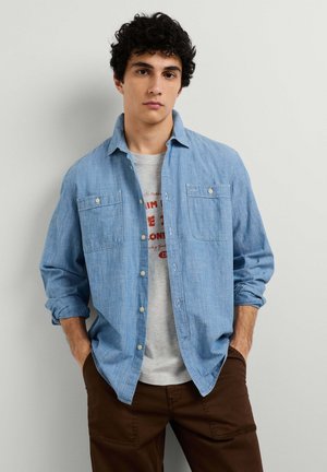 Jonge man met krullend donker haar, die een lichtblauw overhemd draagt over een wit bedrukte T-shirt en een bruine broek, staand met handen in de zakken.