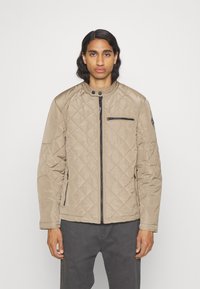 Beige kviltad jacka med ståkrage, dragkedjestängning framtill och bröstficka. Har strukturerad tyg och svarta detaljer.