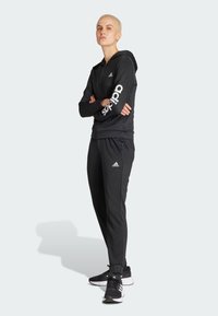 Czarny strój sportowy z zamkiem błyskawicznym, w tym bluza z kapturem z białym logo "adidas" na rękawie oraz dopasowane spodnie, zaprojektowany dla komfortu i swobody ruchów.