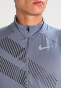 Grå Nike zip-up träningsjacka med hög krage, med svarta horisontella ränder och en liten logotyp på högra bröstpartiet.