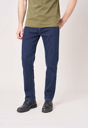 Man draagt donkerblauwe spijkerbroek, zwarte leren laarzen en een olijfgroen overhemd, staand tegen een lichte achtergrond.