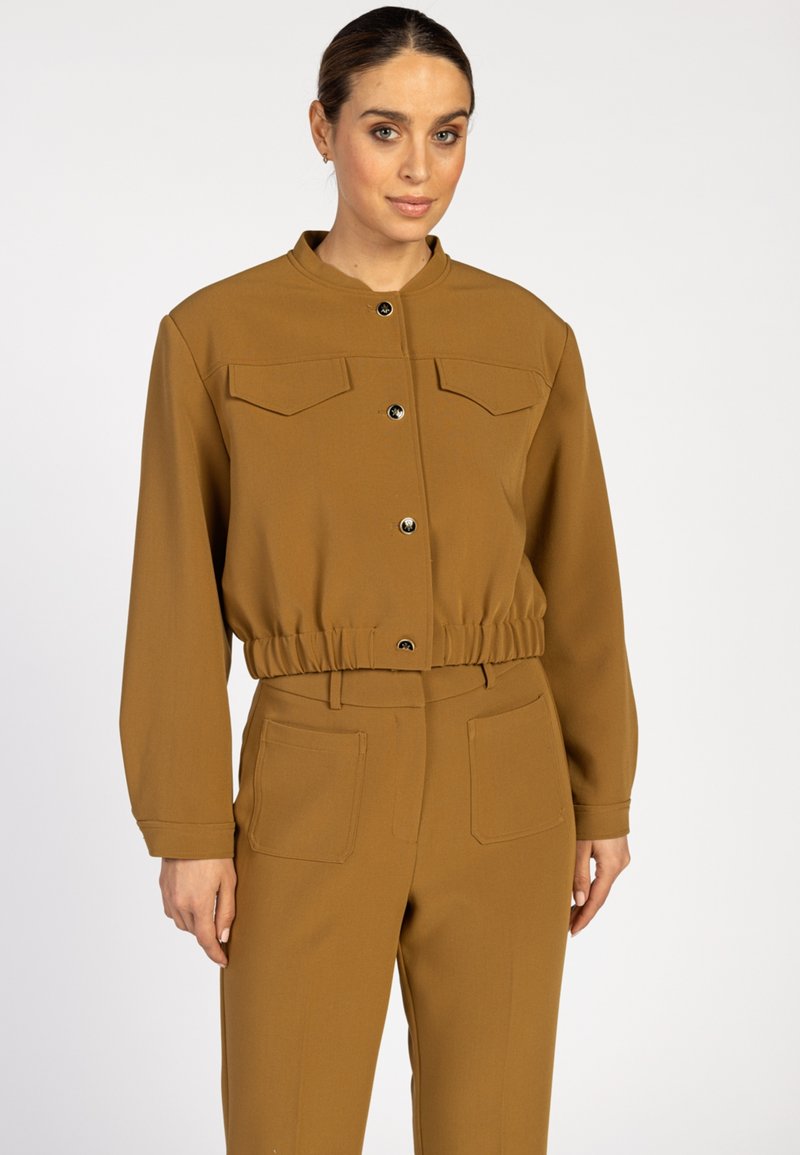 Vrouw in een bruine cropped jas met knopen en bijpassende hooggerokte broek met opgestikte zakken aan de voorkant, staande tegen een witte achtergrond.