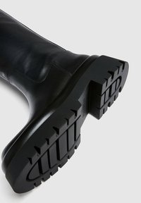 Bota de cuero negro hasta la rodilla con suela de goma gruesa que presenta un patrón de textura y costuras a lo largo del eje.