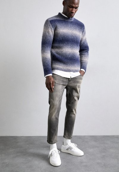 Strikkegenser i gradient blåtoner, høy hals, grå denimbukser, hvite sko, teksturert finish, stående posering mot en nøytral bakgrunn.