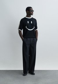 Férfi fekete napszemüvegben, fekete pólóban, melyen nagy fehér mosolygós arc látható, fekete bő szárú nadrágban, szürke fal előtt áll.