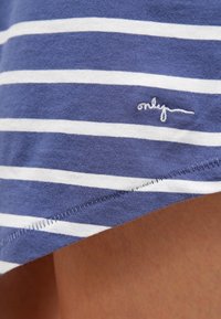 Tissu rayé bleu marine et blanc avec une texture douce. Présente un petit logo brodé en bas et une coupe arrondie au bord inférieur.