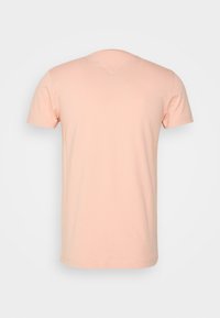 Koszulka z krótkim rękawem w miękkim odcieniu brzoskwini, z okrągłym dekoltem i V-kształtną ozdobą na plecach. Gładka tekstura, minimalistyczny design.