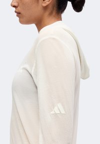 Vue latérale d'une personne portant un haut à capuche beige clair à manches longues avec un logo discret sur le haut du bras.